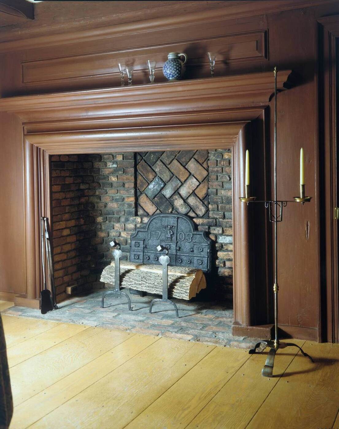 A wide paneled hearth with herringbone brick, iron firedogs, and a candlestand